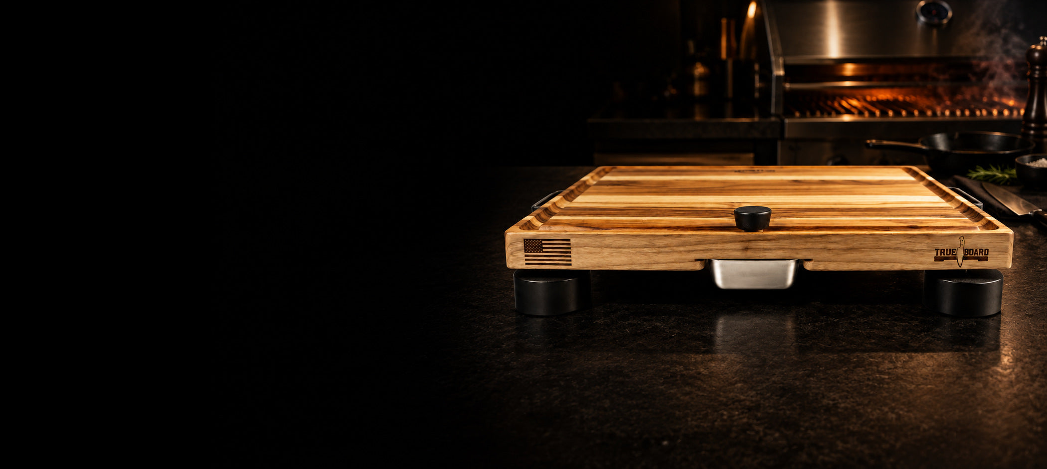 The True Board teak cutting board on a dark stone counter in front of a built-in grill